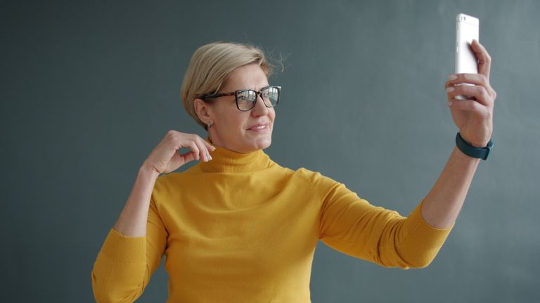 An adult woman in a yellow sweater takes a selfie with her smartphone indoors.