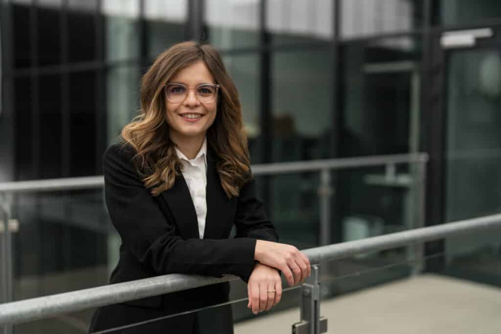 Was anziehen fürs Bewerbungsfoto? 7 Tipps für den perfekten Look 2 Portraibild einer jungen Frau im Businessoutfit. Sie lehnt sich im überkreuzten Armen locker an ein Geländer. - Was anziehen fürs Bewerbungsfoto